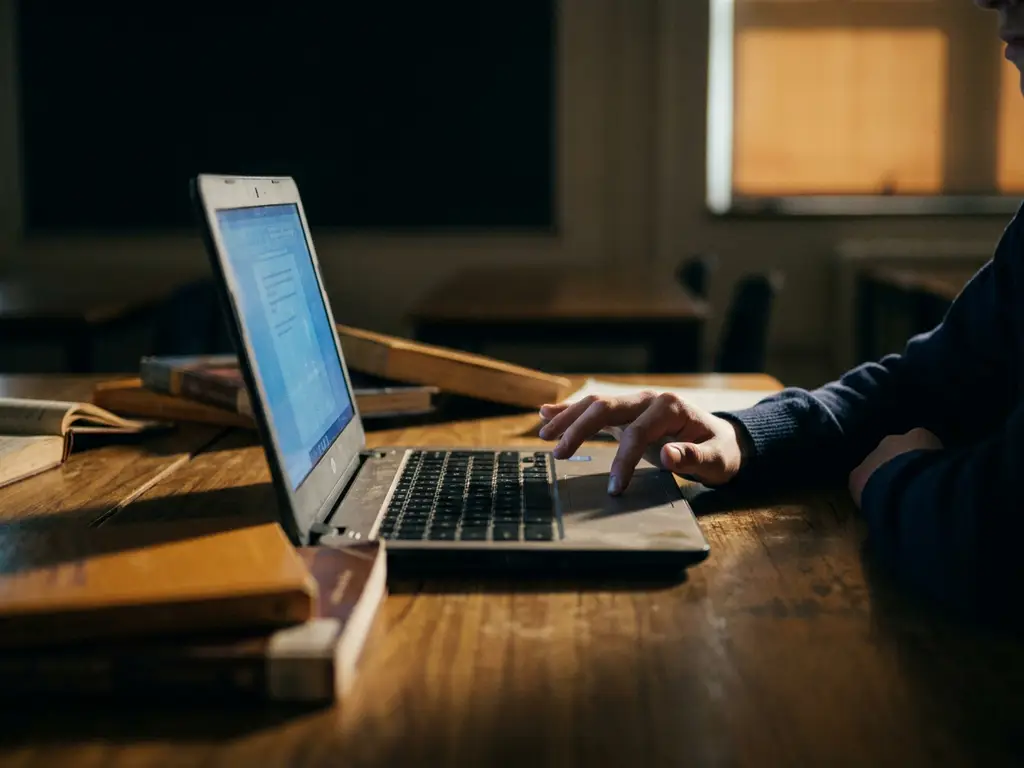 Student scrollt op schoollaptop in donker klaslokaal, blauw schermgloed verlicht hand en versleten houten bureau.